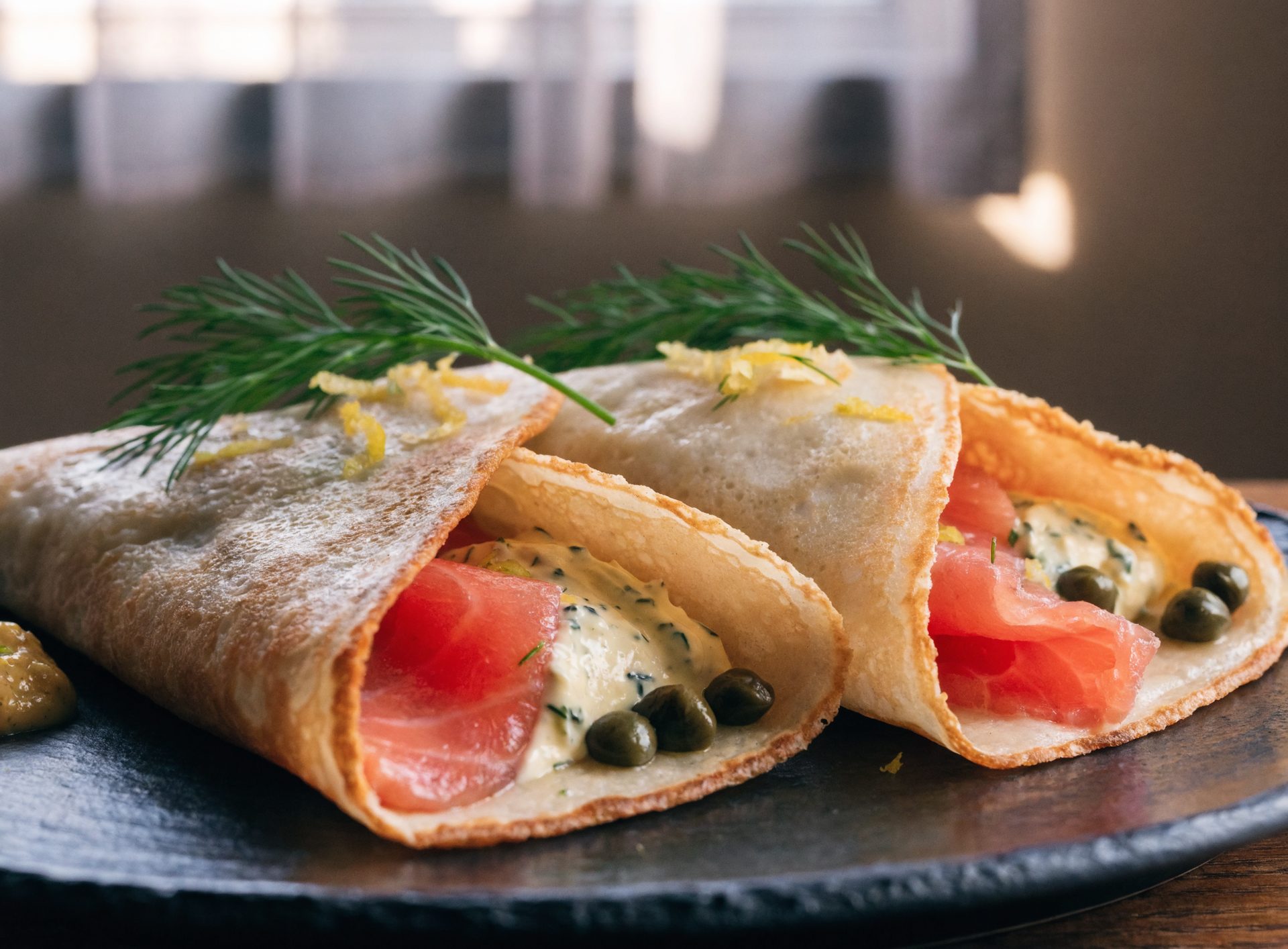 Zwei gefüllte Crêpes mit Lachs und Dill-Senf-Frischkäse auf Schieferplatte, Kapern und Zitrone