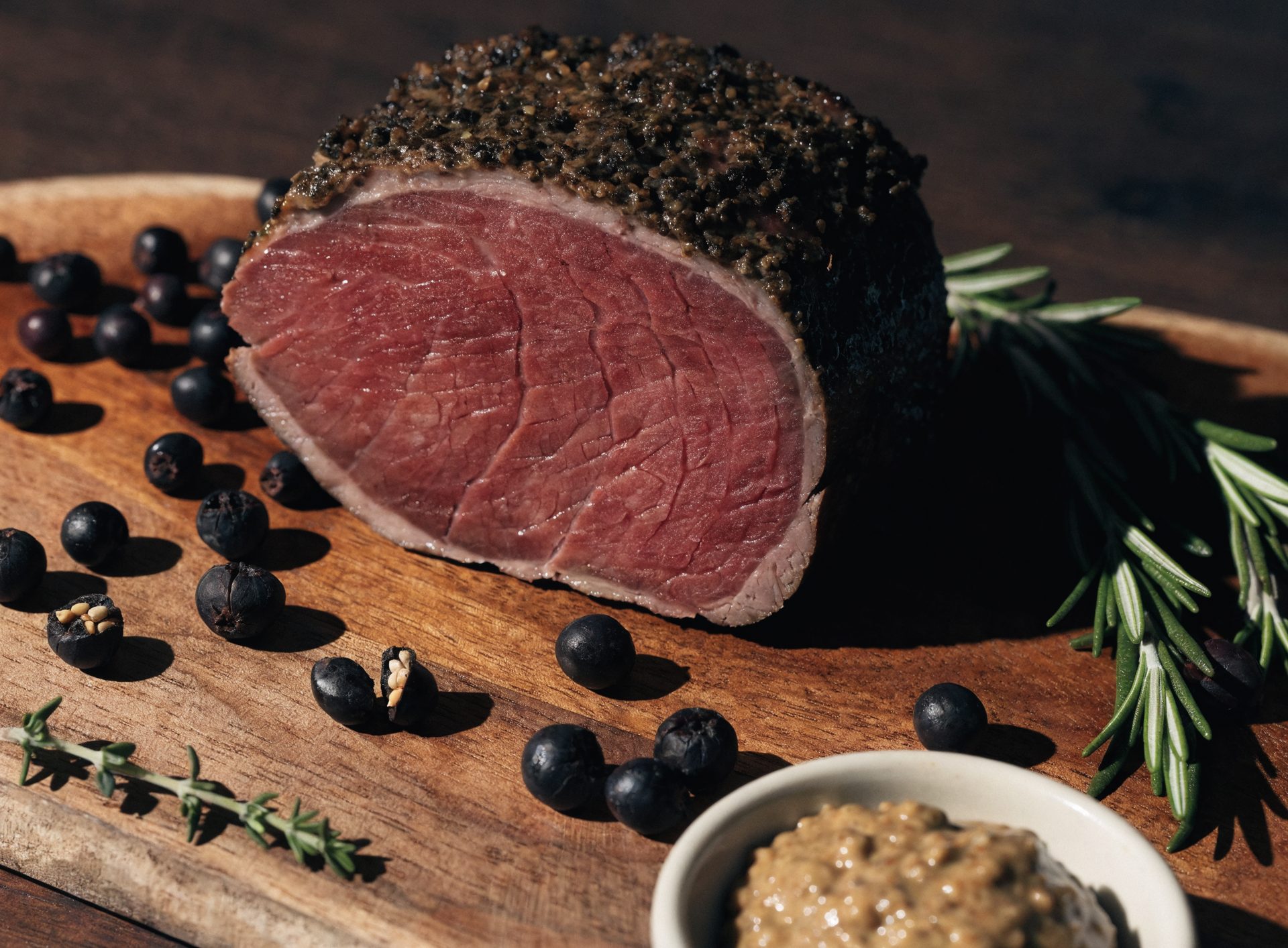 Saftige Scheibe Rehbraten mit dunkler Kräuter-Senf-Kruste auf Holzbrett mit Wacholderbeeren