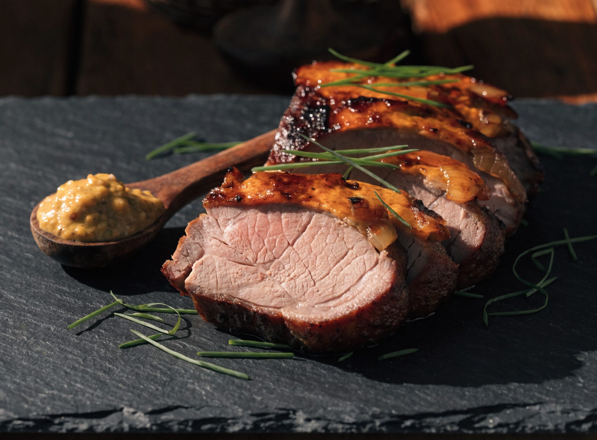 Dicke Scheiben Schweinefilet mit dunkler Zwiebel-Senf-Kruste auf Schieferplatte mit Schnittlauch