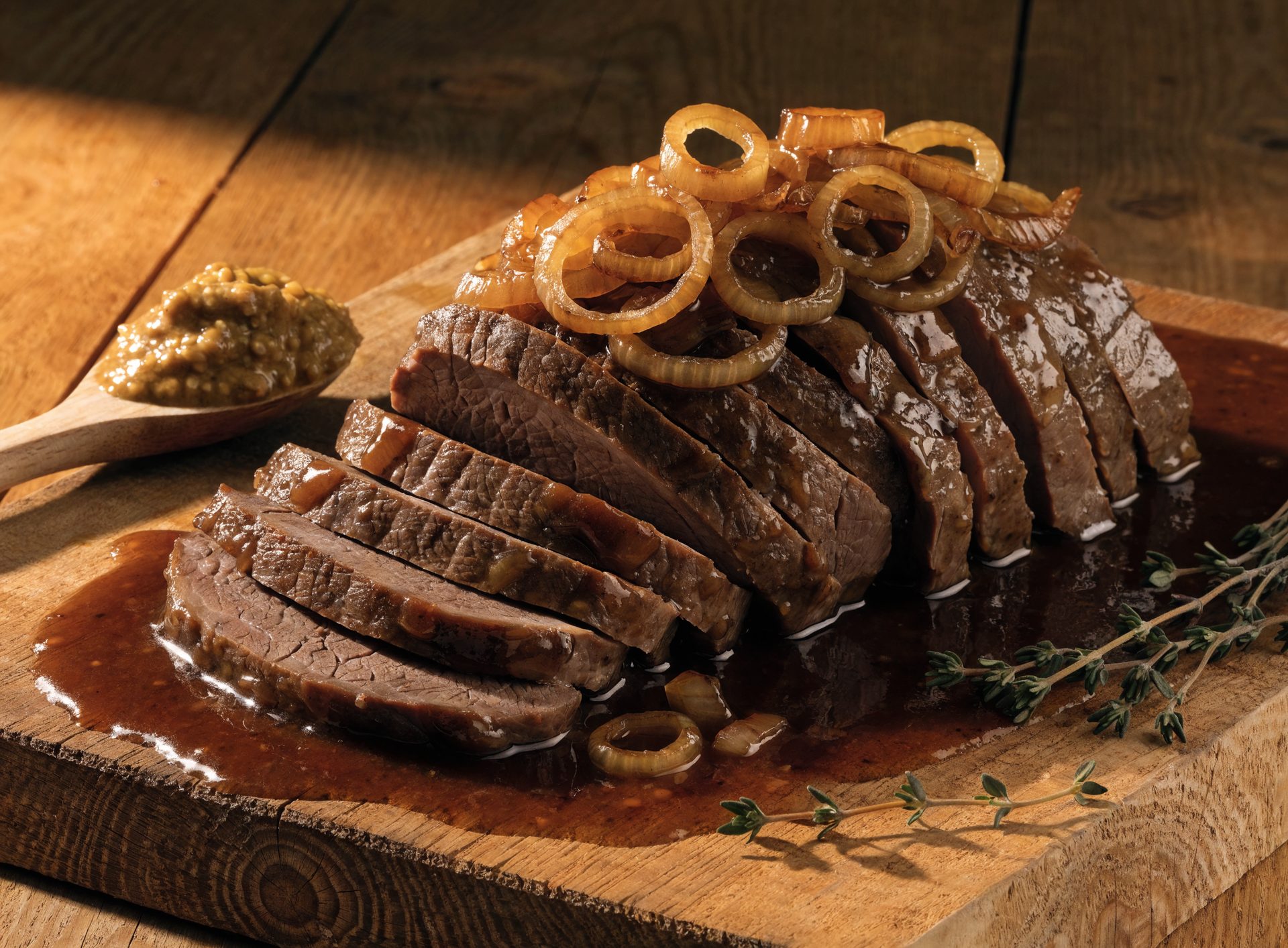 Saftiger Rinderbraten mit karamellisierten Zwiebelringen und dunkler Senf-Bier-Sauce auf Holzbrett